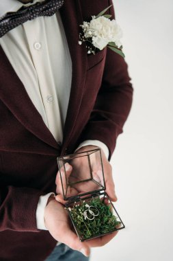 partial view of groom in suit with buttonhole and wedding rings in box in hands clipart
