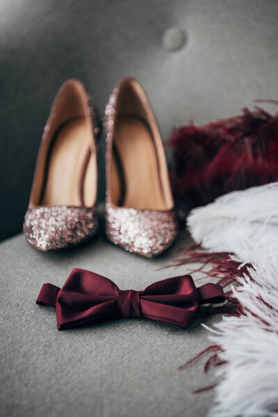 selective focus of bridal shoes, grooms bow tie and feathers for rustic wedding on armchair