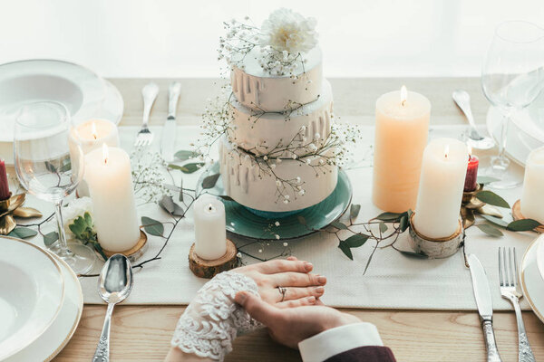 partial view of newlyweds holding hands while sitting at served table with wedding cake, rustic wedding concept