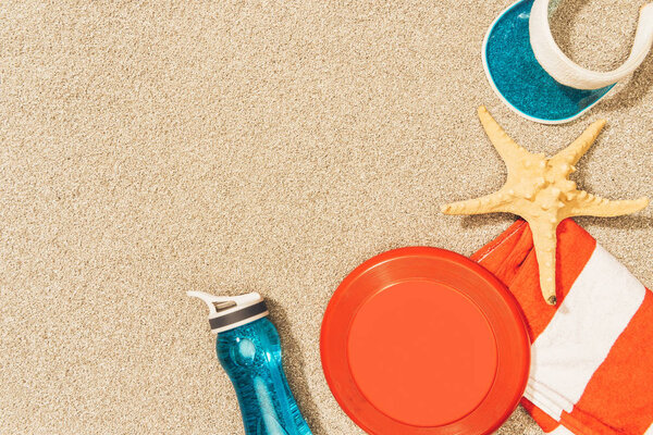 flat lay with water bottle, towel, cap, frisbee and sea star on sand
