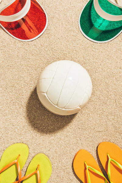 flat lay with white volleyball ball, colorful caps and flip flops arranged on sand