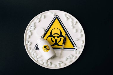Top view of pills, syringe and jar on plate with biohazard sign isolated on black clipart