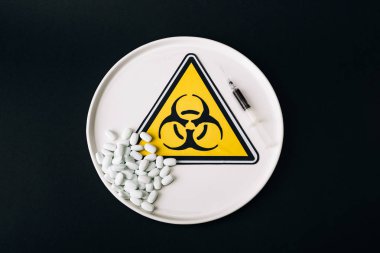 Top view of pills and syringe on plate with biohazard sign isolated on black clipart