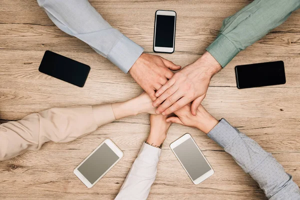 Top view of business partners holding hands by smartphones on table, cropped view