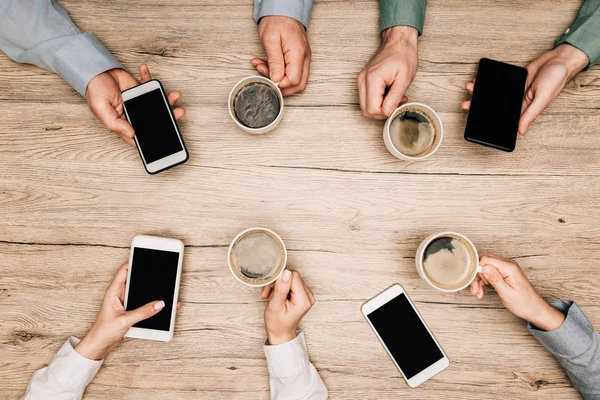 Top view of business people drinking coffee and using smartphones at wooden table, cropped view