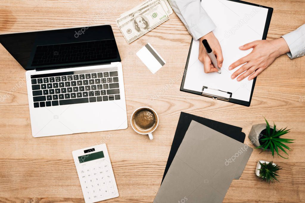 Top view of businessman writing on clipboard with laptop, money and credit card on table