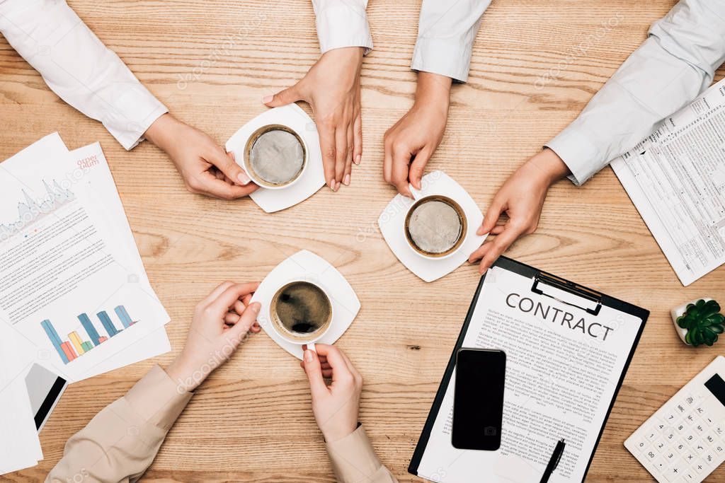 Top view of business people near cups with coffee and paperwork on wooden table, cropped view