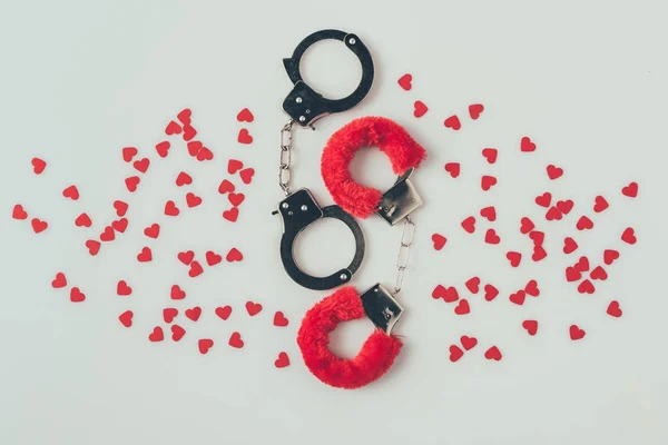 Vue du haut des menottes noires et rouges avec des coeurs de papier éparpillés isolés sur blanc, concept de Saint-Valentin — Photo de stock