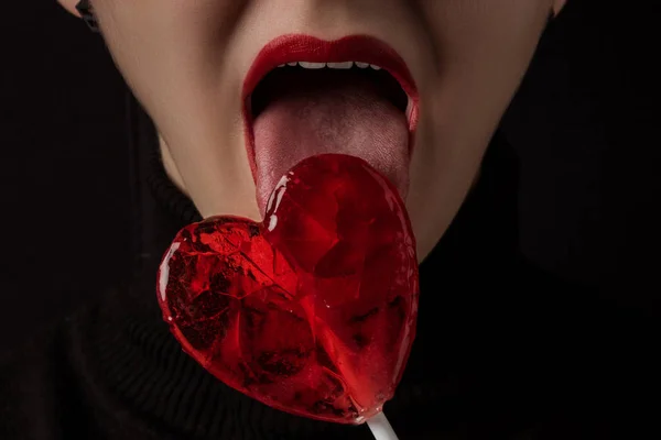 Image recadrée de femme léchant sucette en forme de coeur isolé sur noir, concept de Saint-Valentin — Photo de stock