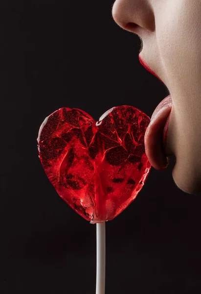 Immagine ritagliata di donna leccare lecca lecca-lecca a forma di cuore rosso isolato su nero, concetto di giorno di San Valentino — Foto stock