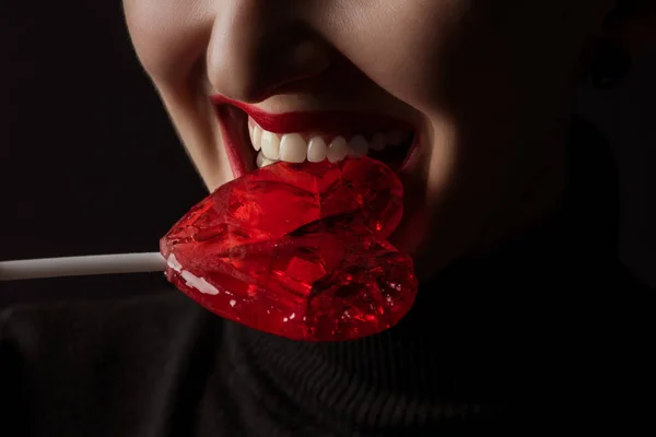 Imagen recortada de mujer mordiendo paleta en forma de corazón aislado en negro, concepto de día de San Valentín - foto de stock