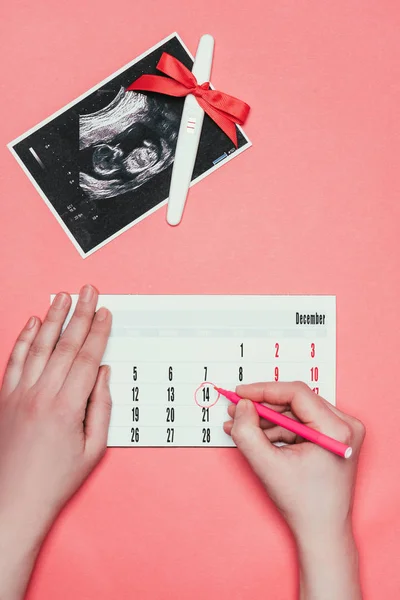 Partial view of woman making note on calendar, ultrasound scan and pregnancy test with ribbon isolated on pink — Stock Photo