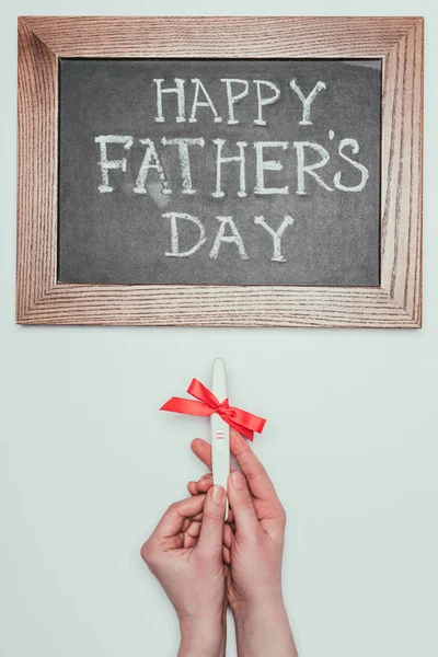 Cropped shot of woman holding pregnancy test with ribbon and happy fathers day lettering on blackboard isolated on grey — Stock Photo