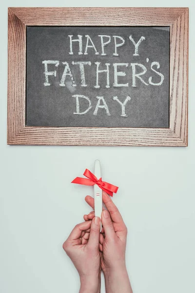 Cliché recadré d'une femme tenant un test de grossesse avec un ruban et un lettrage heureux de la fête des pères sur un tableau noir isolé sur gris — Photo de stock