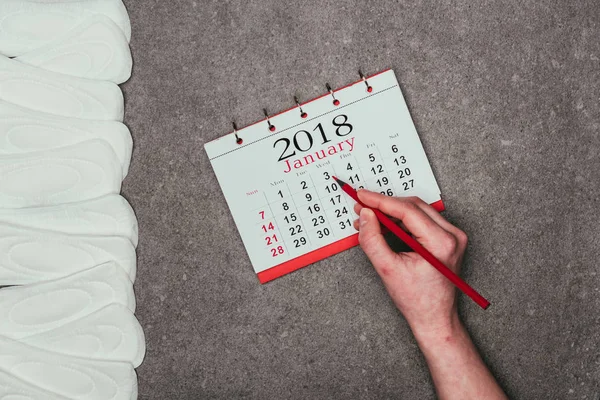 Tiro recortado de mulher apontando na data no calendário com almofadas menstruais em torno de superfície cinzenta — Fotografia de Stock