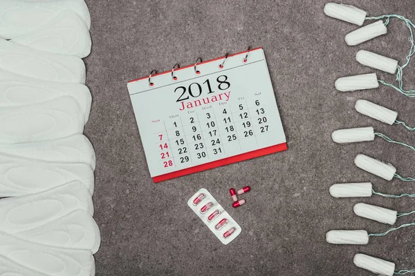 Vista superior de almohadillas menstruales dispuestas y tampones, calendario y pastillas en la superficie gris - foto de stock