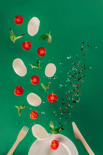 Vista de perto de queijo mussarela, tomate cereja, espinafre e especiarias caindo na placa isolada em verde — Fotografia de Stock