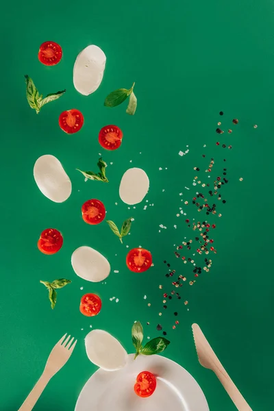 Close up view of mozzarella cheese, cherry tomatoes, spinach and spices falling on plate isolated on green — Stock Photo