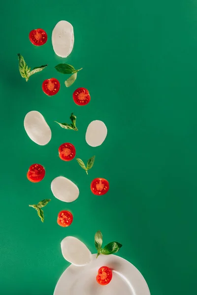 Close up view of mozzarella cheese, cherry tomatoes, spinach falling on plate isolated on green — Stock Photo