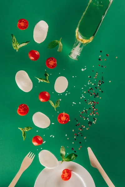 Close up view of mozzarella cheese, cherry tomatoes, spinach, spices and olive oil falling on plate isolated on green — Stock Photo