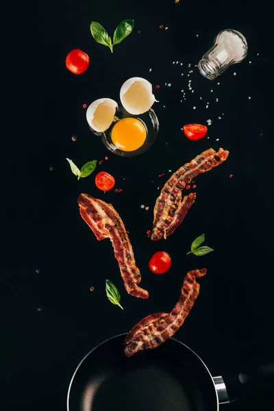 Vista da vicino di pezzi di pancetta, pomodorini e tuorlo d'uovo crudo che cadono sulla padella isolata sul nero — Foto stock