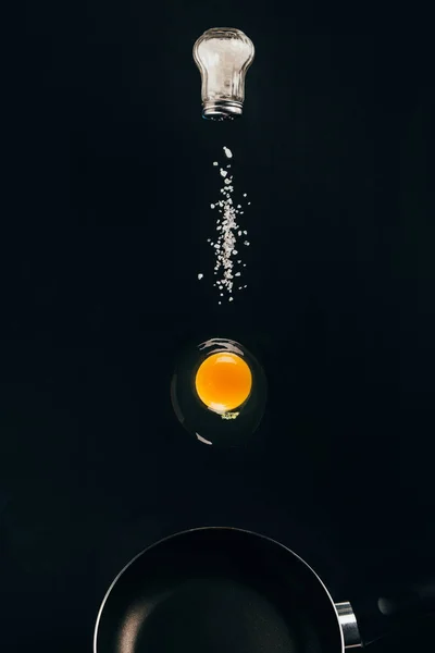 Close up view of raw egg yolk and salt falling on frying pan isolated on black — Stock Photo