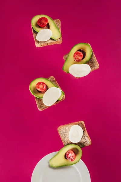 Vista de perto de torradas, pedaços de abacate, queijo mussarela e tomate cereja caindo no prato isolado em rosa — Fotografia de Stock