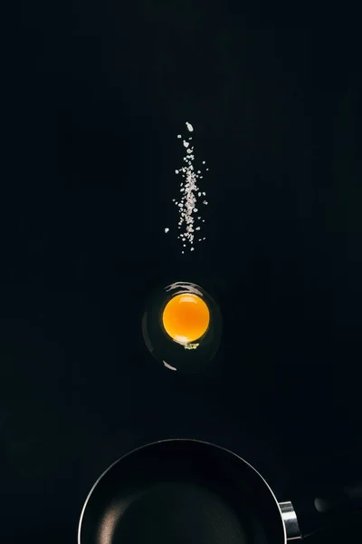 Close up view of raw egg yolk and salt falling on frying pan isolated on black — Stock Photo