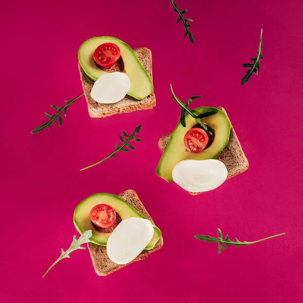 Vista de cerca de tostadas levitantes, trozos de aguacate, tomates cherry, queso mozzarella y rúcula aislados en rosa - foto de stock