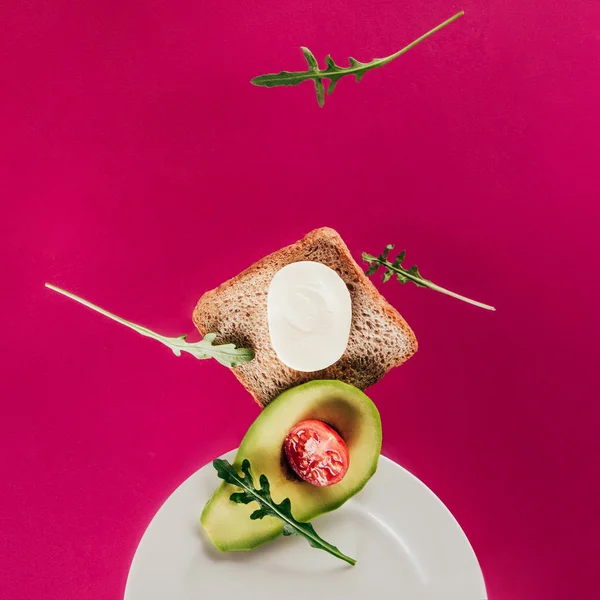 Vista de perto de torradas, pedaços de abacate, tomate cereja, queijo mussarela e arugula caindo no prato isolado em rosa — Fotografia de Stock