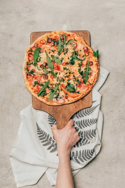 Close-up view of hand holding pizza on wooden cutting board on light background — Stock Photo