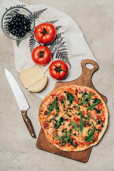 Pizza entera sobre tabla de cortar de madera con tomates, queso y aceitunas sobre fondo claro - foto de stock