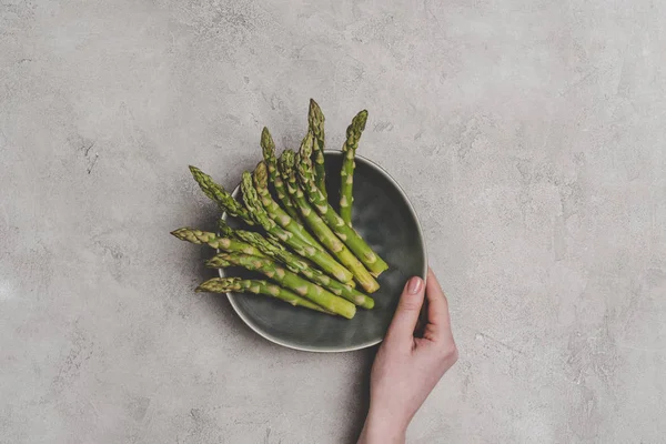 Partial top view of person holding plate with fresh healthy asparagus on grey — Stock Photo