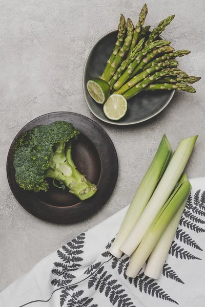 Vista superior de verduras verdes frescas y saludables en platos y servilleta en gris - foto de stock