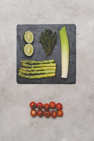 Draufsicht auf Tomaten, Limetten, Lauch und Spargel auf Schiefertafel auf grau — Stockfoto