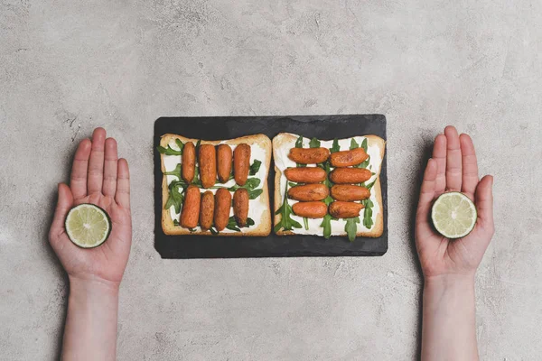 Cropped shot of human hands with  slices of lime and healthy sandwiches with arugula and baby carrots on slate board — Stock Photo