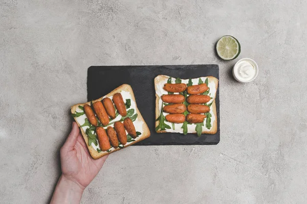 Partial top view of human hand and healthy sandwiches with arugula and baby carrots — Stock Photo