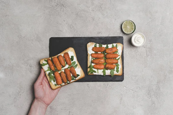 Vista superior parcial da mão humana e sanduíches saudáveis com arugula e cenouras de bebê — Fotografia de Stock