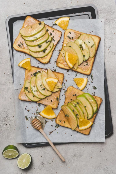 Top view of healthy sandwiches with slices of apple and orange, lime and honey dipper on grey — Stock Photo