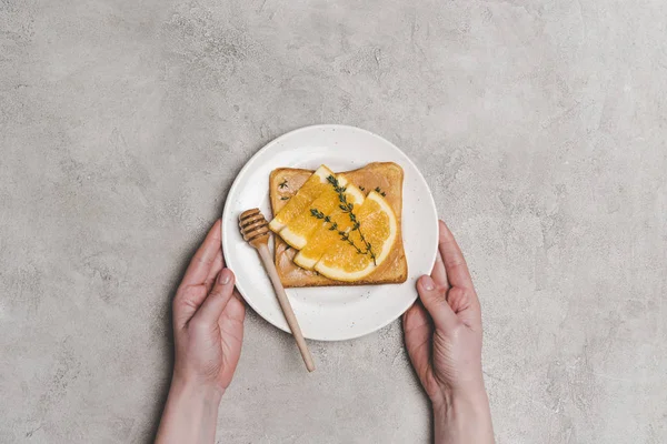 Vista superior parcial da pessoa segurando prato com sanduíche saudável fresco com mel e fatias de laranja no cinza — Fotografia de Stock