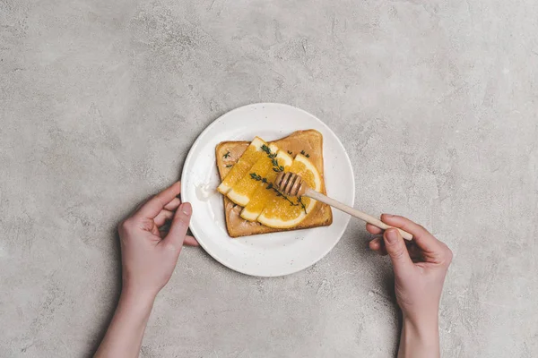 Vista superior de mãos humanas com mel dipper e sanduíche saudável com fatias de laranja no cinza — Fotografia de Stock