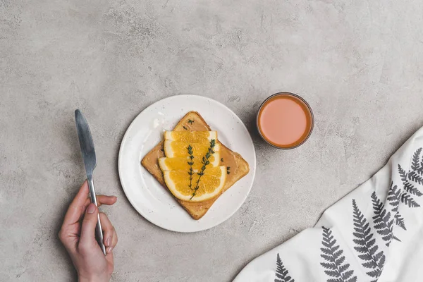 Vue partielle du dessus de la personne tenant le couteau, verre de jus et pain grillé avec des tranches d'orange sur gris — Photo de stock