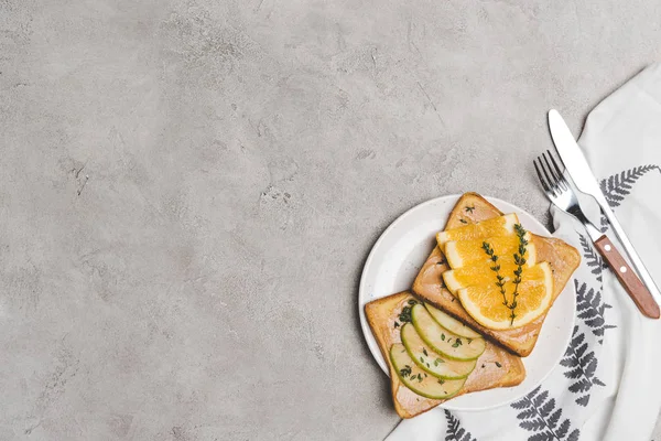 Top view of healthy toasts with apple and orange slices on plate with fork and knife on grey — Stock Photo