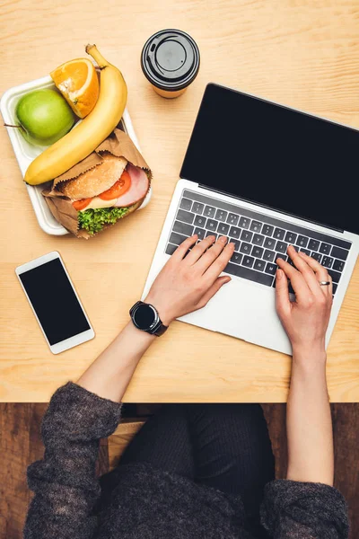 Imagen recortada de la mujer que usa el ordenador portátil en la mesa con comida en el contenedor — Stock Photo