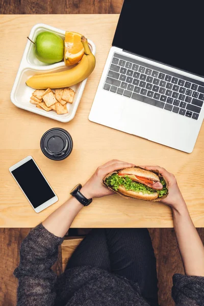 Imagem cortada de mulher comendo hambúrguer à mesa com frutas e gadgets — Fotografia de Stock