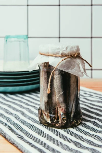 Montón de pescado salado en frasco envuelto por cuerda en toalla con platos y vidrio detrás - foto de stock