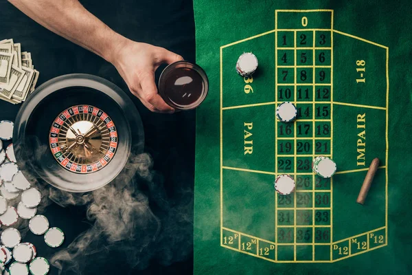 Hand with whiskey in glass by casino table with bets and roulette — Stock Photo