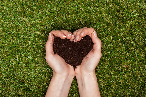 Imagem cortada de mulher segurando o solo em forma de coração em mãos acima da grama verde, conceito de dia de terra — Fotografia de Stock