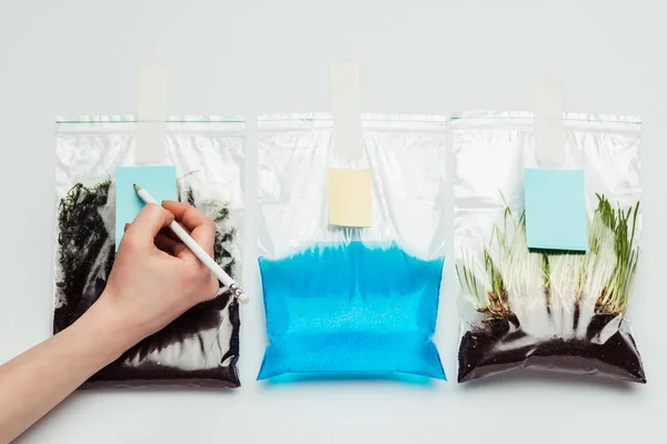 Обрезанный образ женщины подписания пластиковые пакеты с почвой, водой и саженцы изолированы на белом, земля день концепции — стоковое фото