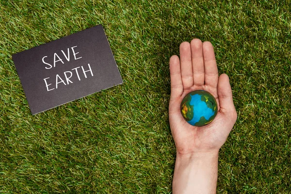 Imagen recortada del hombre sosteniendo el modelo de tierra en la mano con signo salvar la tierra en la hierba verde, concepto de día de la tierra - foto de stock